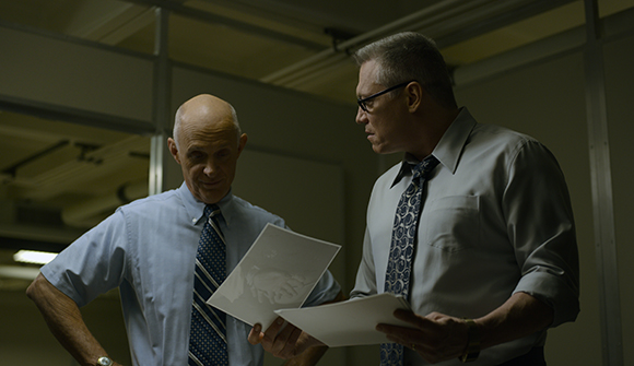 Two men in button-down shirts and ties examine documents in a dimly lit room.