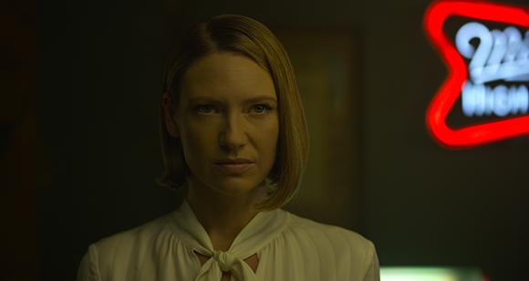 A woman with blonde hair and a white blouse looks forward in a dimly lit setting.