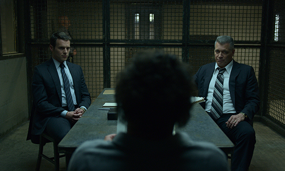 Two men in suits sit at a table in a room with metal mesh walls, facing a person in the foreground.