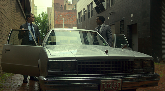 Two men in suits stand by a silver vintage car with open doors in a narrow city alley.