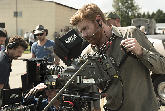 A man with a beard looks through the viewfinder of a large RED cinema camera on a film set.