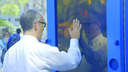 A man in a white dress shirt reaches out to touch a blue glass window, seeing his reflection.