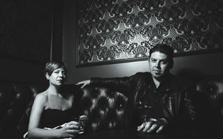 A man and a woman sit on a tufted leather booth in a dimly lit room with patterned wallpaper.