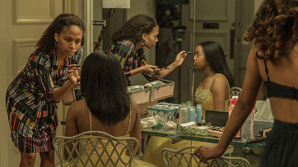 A makeup artist in a patterned dress applies makeup to a woman seated in front of a mirror.
