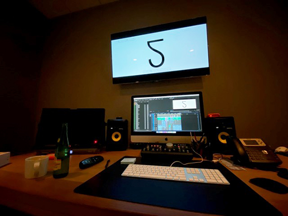 A dark studio desk with a computer monitor, large screen displaying a large number 5, speakers, and a keyboard.