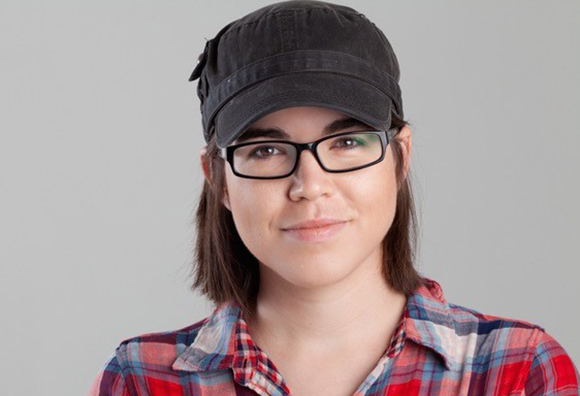 A person with short brown hair wearing a black cadet cap, black-rimmed glasses, and a red plaid shirt.
