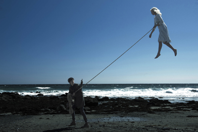 A person in a white dress floats in the sky, held by a rope by a person standing on a dark beach.