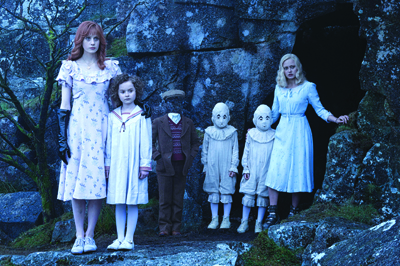Six people in vintage attire stand in a line before a dark cave entrance in a blue-toned forest.