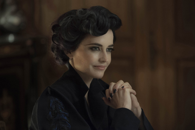 A woman with dark wavy hair and a black high-collared outfit sits with her hands clasped.