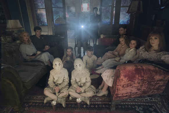 A group of people sit in a dark, vintage living room with two figures in white masks sitting in the foreground.