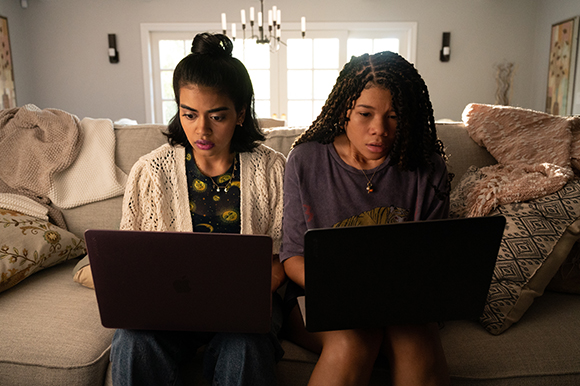 Two young women sit side-by-side on a couch, each focused on their own open laptop.
