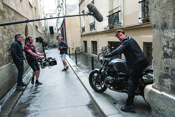 A film crew with a camera and boom mic films a man riding a black motorcycle through a narrow city alley.