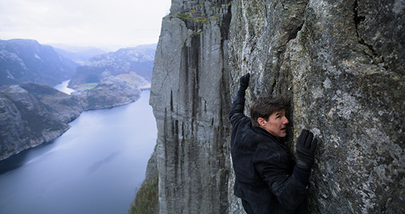 A man in dark clothing climbs a vertical grey rock cliff above a large body of water.