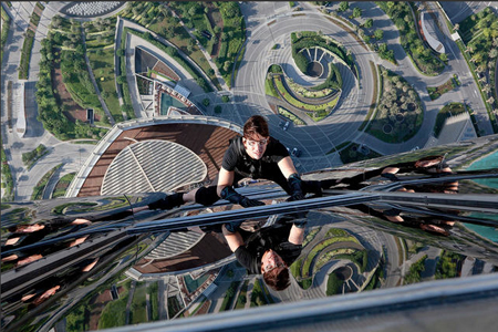 High-angle view of a person in black gear climbing a reflective glass building overlooking a city park.