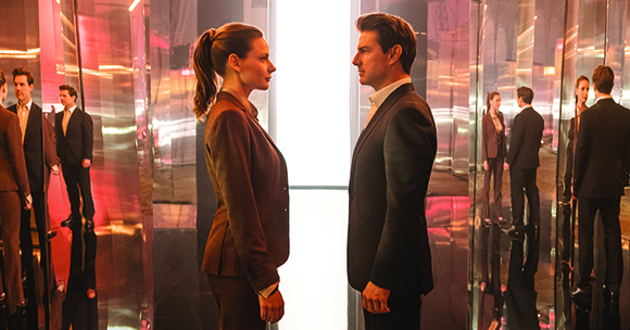 A man and woman in suits stand face-to-face in a room filled with reflective surfaces.