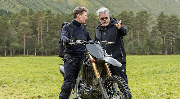 A man on a dirt bike listens to another man pointing toward the distance in a grassy field.
