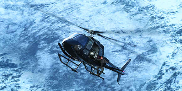 A black helicopter flies over a vast, snow-covered mountain range under a bright sky.