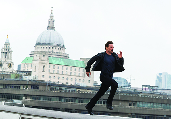 A man in dark clothing runs across a rooftop with a large domed cathedral in the background.