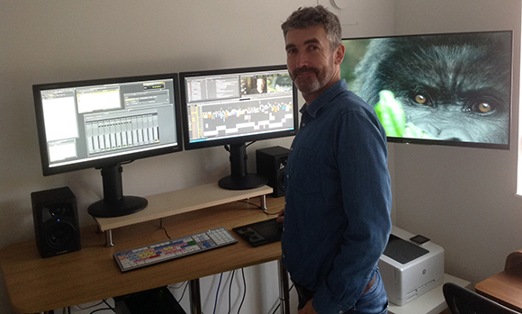 A man in a blue shirt stands in a studio with two computer monitors and a large screen showing a gorilla's face.