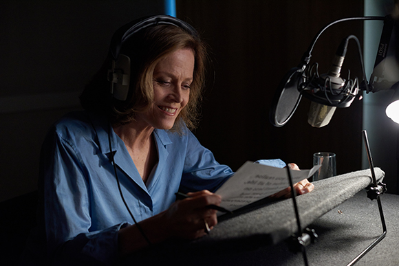 A woman in a blue shirt reads a script while wearing headphones in a dark recording studio.