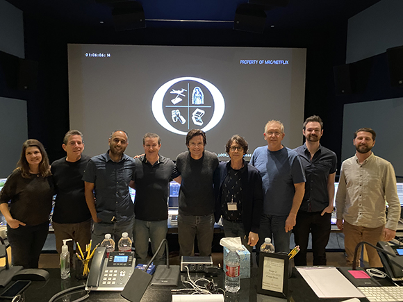 Seven people stand in a row in a dark room before a large screen displaying a circular graphic.