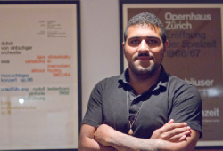 A man with dark hair and a beard stands with arms crossed in front of two framed posters.