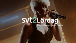 A blonde singer holds a microphone and performs under stage lighting.