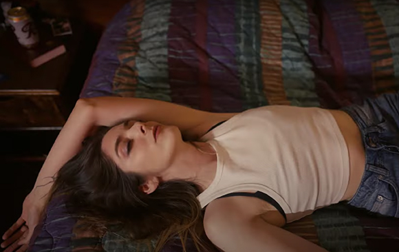 A woman in a white tank top and denim shorts lies on a striped bedspread with her eyes closed.
