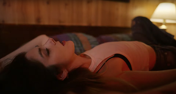 A person with dark hair lies on a bed in a warm, dimly lit room.