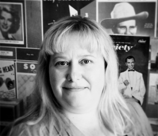 Black and white portrait of a blonde woman smiling in front of various magazine covers.