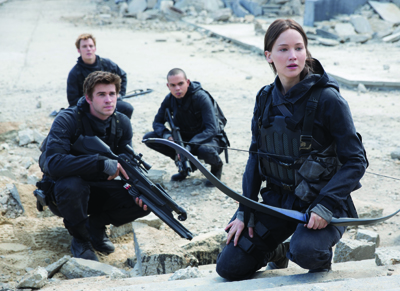 Four people in black tactical clothing crouch among concrete debris, one holding a bow and one holding a rifle.