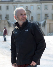 A man with graying hair and a beard wearing a black North Face jacket stands in front of a large building.