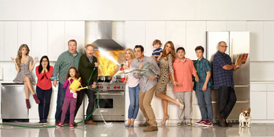 A large group of people posing in a white kitchen with a fire burning in the stove.