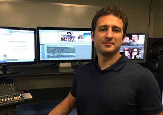A man in a dark polo shirt stands in front of several computer monitors in a studio setting.