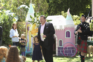A man in a dark suit stands near a pink playhouse while a bagpiper performs in a kilt.