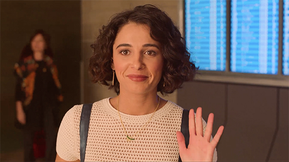 A woman with curly dark hair smiles and raises her hand in a wave.
