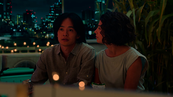 A man and a woman sit side-by-side outdoors at night with city lights blurred in the background.