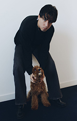 A person in black clothing crouches next to a small, curly-haired brown dog against a white wall.