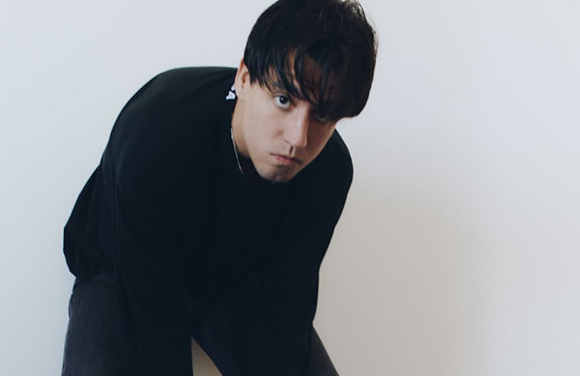 A man with dark hair wearing a black long-sleeved shirt, leaning forward against a white background.