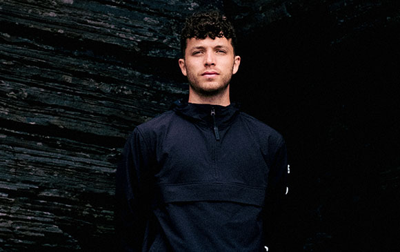 A man with curly hair wearing a dark navy quarter-zip pullover stands before a dark, textured rock wall.