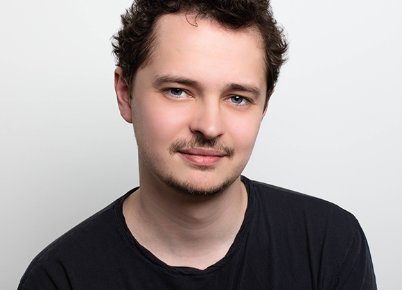 A young man with short brown hair and a light beard wearing a black t-shirt against a white background.