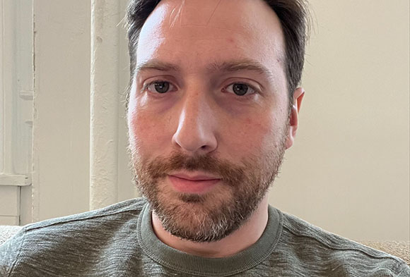 A man with short brown hair and a beard wearing a green crewneck shirt.