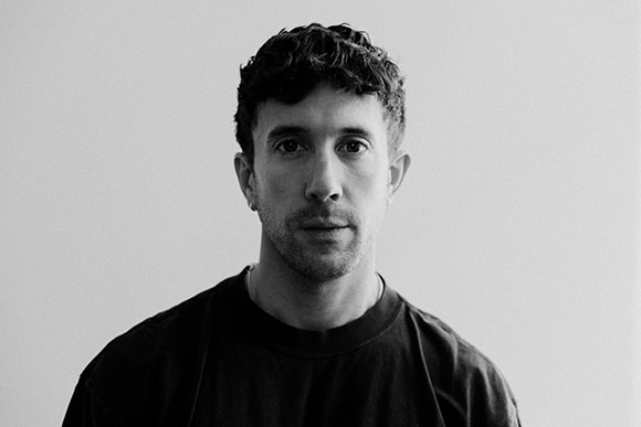Black and white headshot of a man with short, wavy hair wearing a dark t-shirt against a plain background.