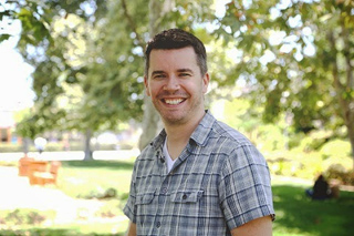 A smiling man with short dark hair wearing a grey plaid shirt stands in a sunlit park.