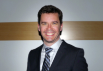 A smiling man wearing a dark suit, light blue shirt, and striped tie against a neutral wall.