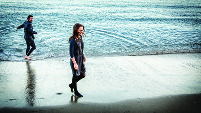 A woman in a dark dress walks on a sandy beach while a man stands in the shallow water behind her.