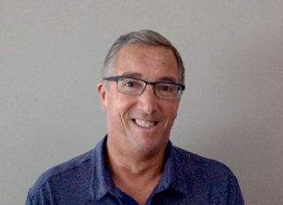 A smiling middle-aged man with short graying hair and glasses wearing a blue button-down shirt.