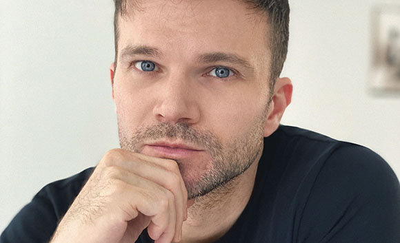 Close-up portrait of a man with short brown hair and blue eyes resting his chin on his hand.