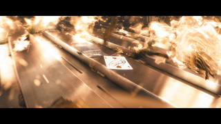 A single playing card rests on a metallic surface surrounded by flames and flying sparks.