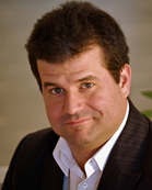 A man with dark hair wearing a dark pinstripe suit and a white collared shirt.